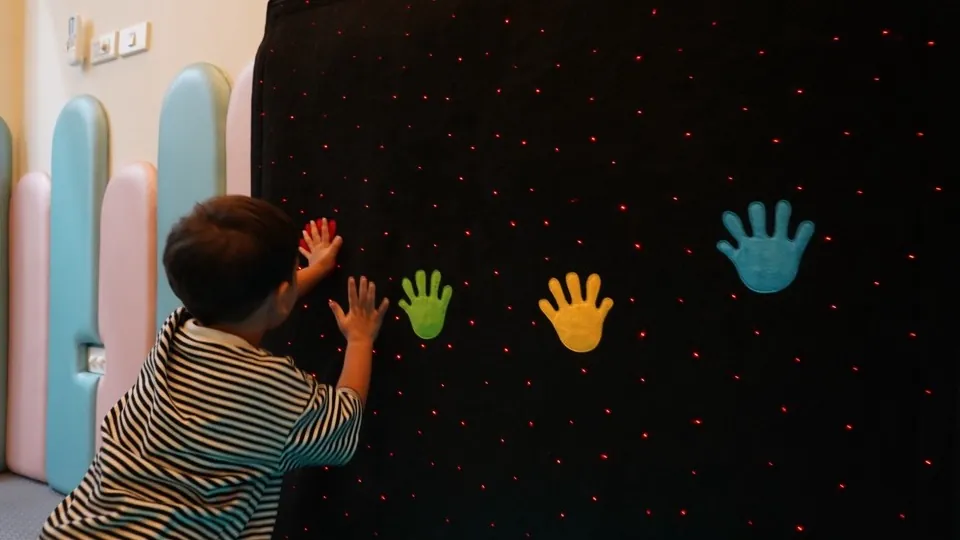เด็กชายแตะแผ่นรูปมือหลากสีบนผนังไฟในห้อง Multisensory Room ที่ InterRehab NeuroBridge